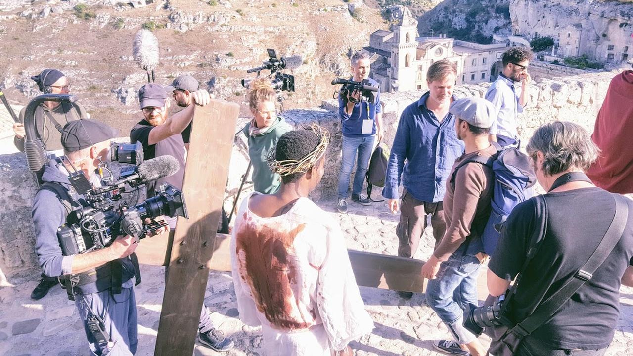 Bei Dreharbeiten zum Filmprojekt "Das Neue Evangelium" von Milo Rau steht Jesusdarsteller Yvan Sagnet auf einer steilen Felsentreppe im blutigen Gewand und Dornenkrone mit dem Rücken zur Kamera. Um ihn herum stehen Milo Rau und das Kamerateam.
