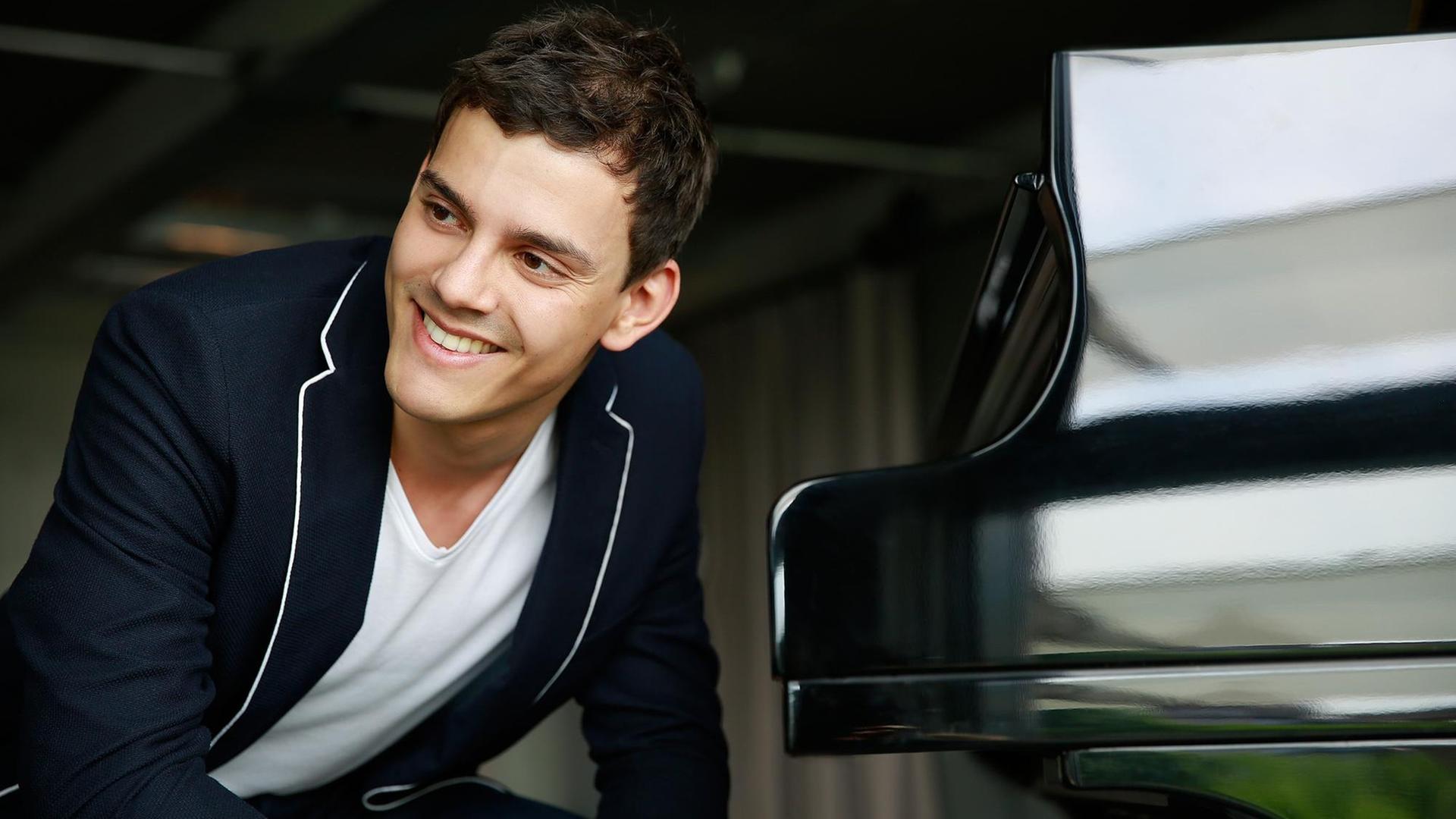 Der Pianist sitzt und schaut rechts seitlich in die Kamera, er trägt ein weißes t-shirt und ein blaues Jacket. Von ihm links aus steht der schwarze glänzende Flügel, zugeklappt. Der Pianist sitzt und schaut rechts seitlich in die Kamera, er trägt ein weißes t-shirt und ein blaues Jacket. Von ihm links aus steht der schwarze glänzende Flügel, zugeklappt.