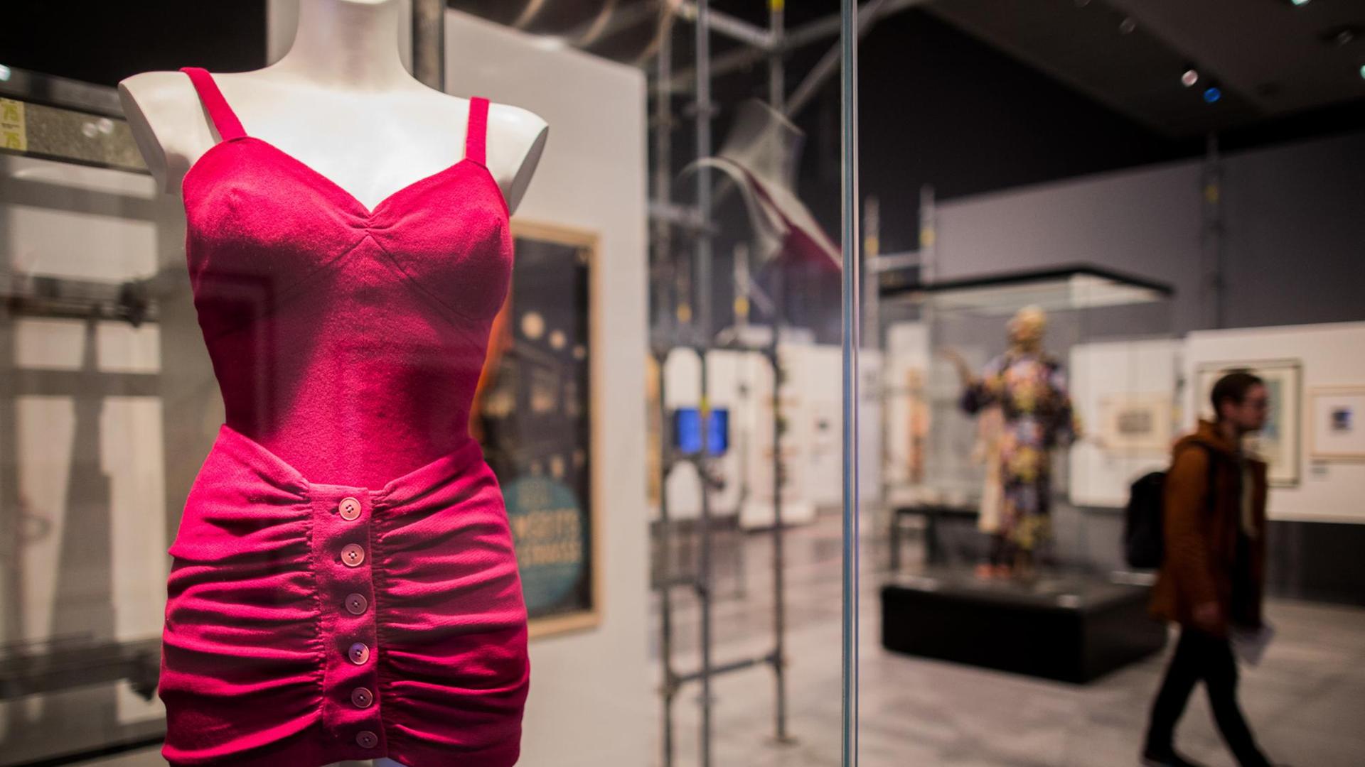 In einem Schaukasten ist ein roter Badeanzug zu sehen, im Hintergrund geht ein Mann durch die Ausstellung In einem Schaukasten ist ein roter Badeanzug zu sehen, im Hintergrund geht ein Mann durch die Ausstellung