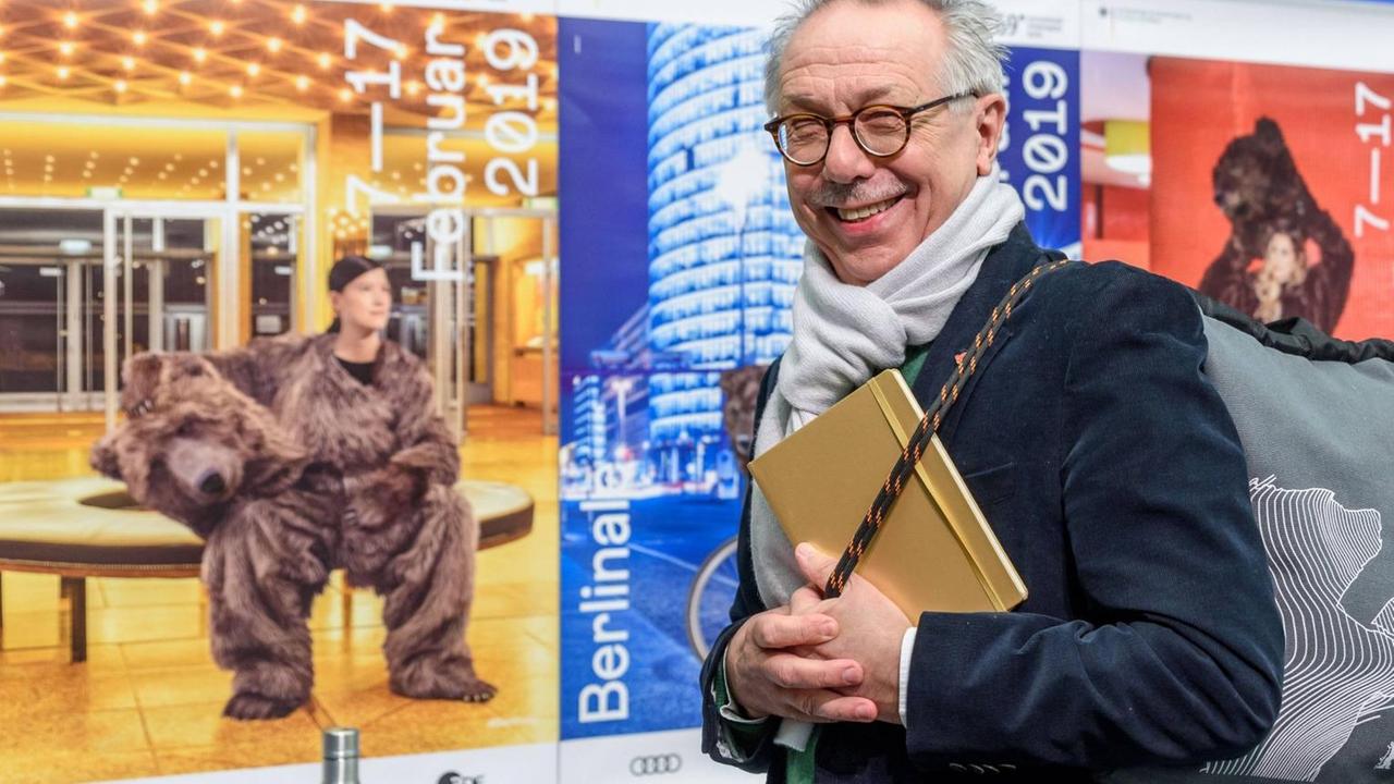 Dieter Kosslick verlässt das Podium der Pressekonferenz und blickt freundlich in die Kamera, während er eine graue Berlinaletasche schultert. Dieter Kosslick verlässt das Podium der Pressekonferenz und blickt freundlich in die Kamera, während er eine graue Berlinaletasche schultert.