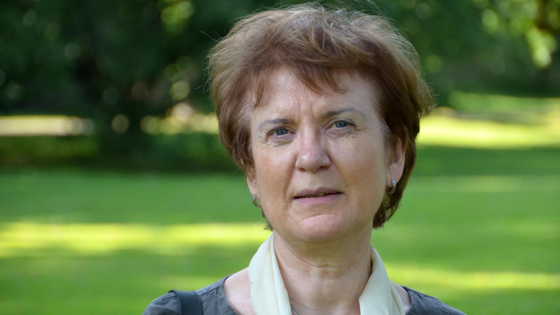 Eine kurzhaarige Frau im mittleren Alter steht auf einer Wiese, im Hintergrund sind Bäume zu sehen. Eine kurzhaarige Frau im mittleren Alter steht auf einer Wiese, im Hintergrund sind Bäume zu sehen.