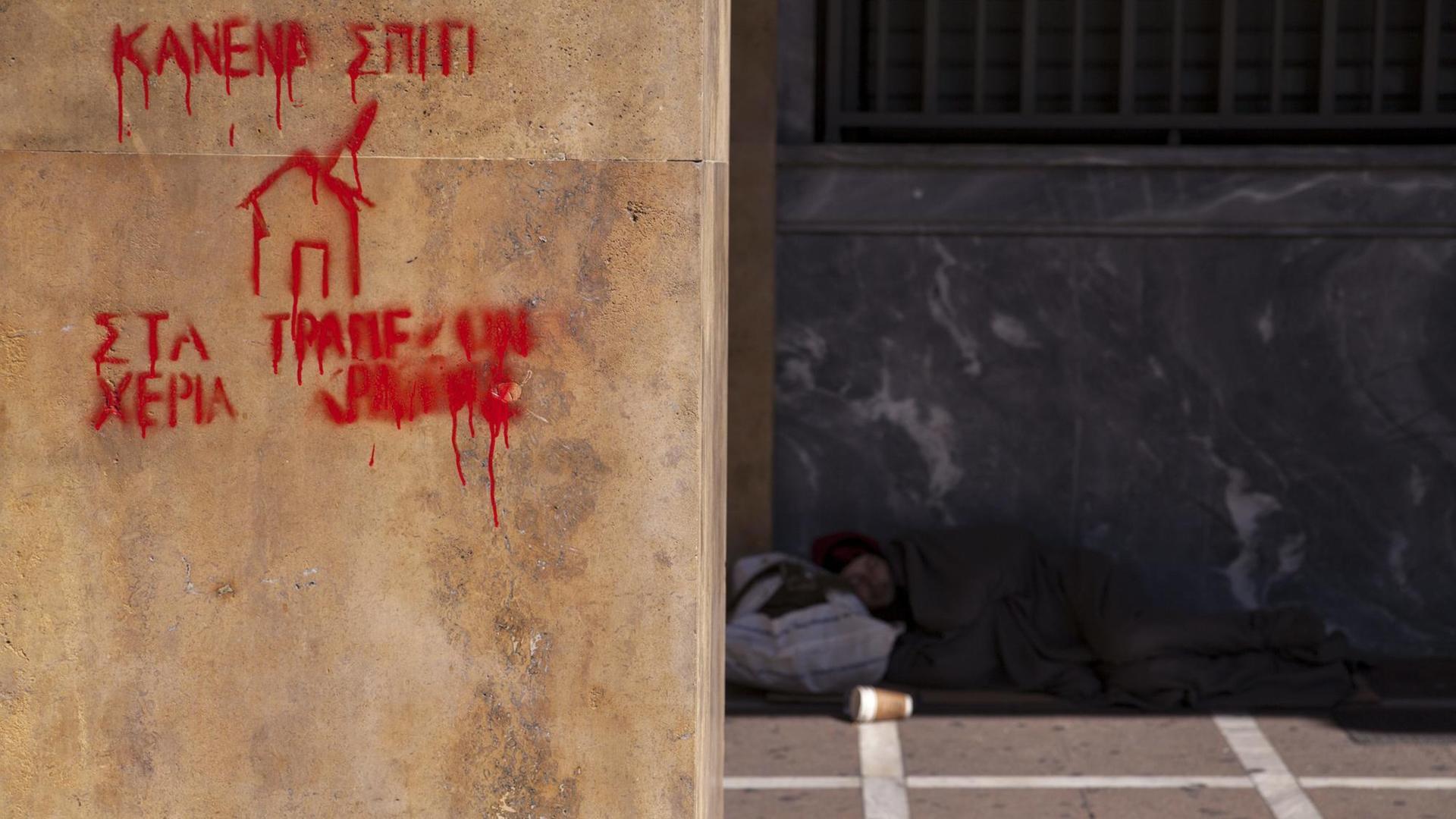 Der Schriftzug "Kein Haus in den Händen eines Bankers" steht in roter Farbe an einer Hauswand, im Hintergrund ist ein Obdachloser zu sehen Der Schriftzug "Kein Haus in den Händen eines Bankers" steht in roter Farbe an einer Hauswand, im Hintergrund ist ein Obdachloser zu sehen