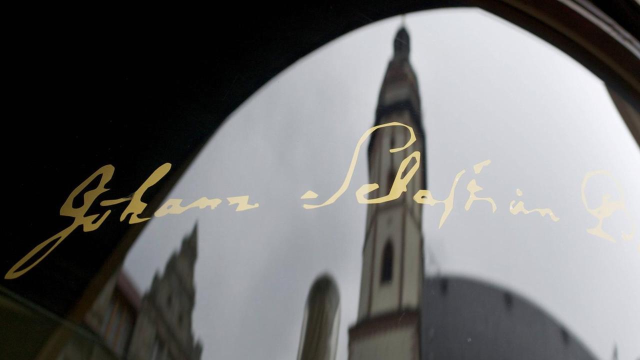 Die Unterschrift des Komponisten Johann Sebastian Bachs in golgener Schrift auf einer Glasscheibe, mit der Spiegelung der Thomaskirche in Leipzig. Die Unterschrift des Komponisten Johann Sebastian Bachs in golgener Schrift auf einer Glasscheibe, mit der Spiegelung der Thomaskirche in Leipzig.