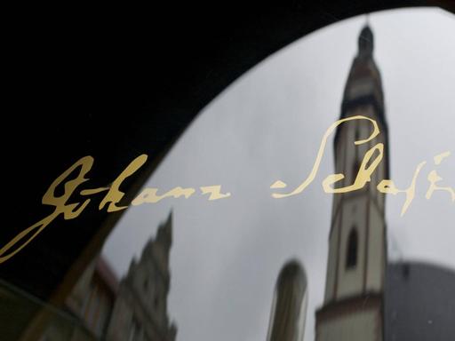 Die Unterschrift des Komponisten Johann Sebastian Bachs in golgener Schrift auf einer Glasscheibe, mit der Spiegelung der Thomaskirche in Leipzig.