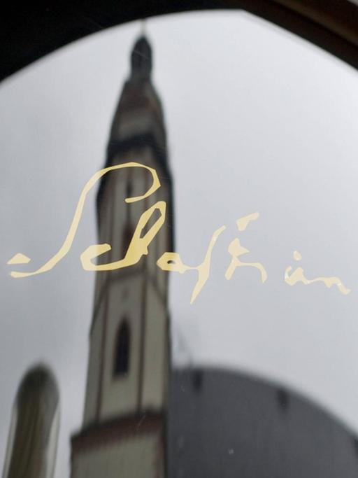 Die Unterschrift des Komponisten Johann Sebastian Bachs in golgener Schrift auf einer Glasscheibe, mit der Spiegelung der Thomaskirche in Leipzig.