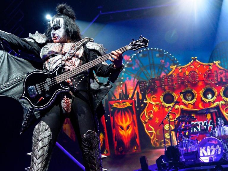 Gene Simmons, der dämonisch geschminkte und verkleidete Bassist der Band Kiss, steht in theatralischer Pose auf einer Bühne, die wie ein Horror-Zirkus aussieht.