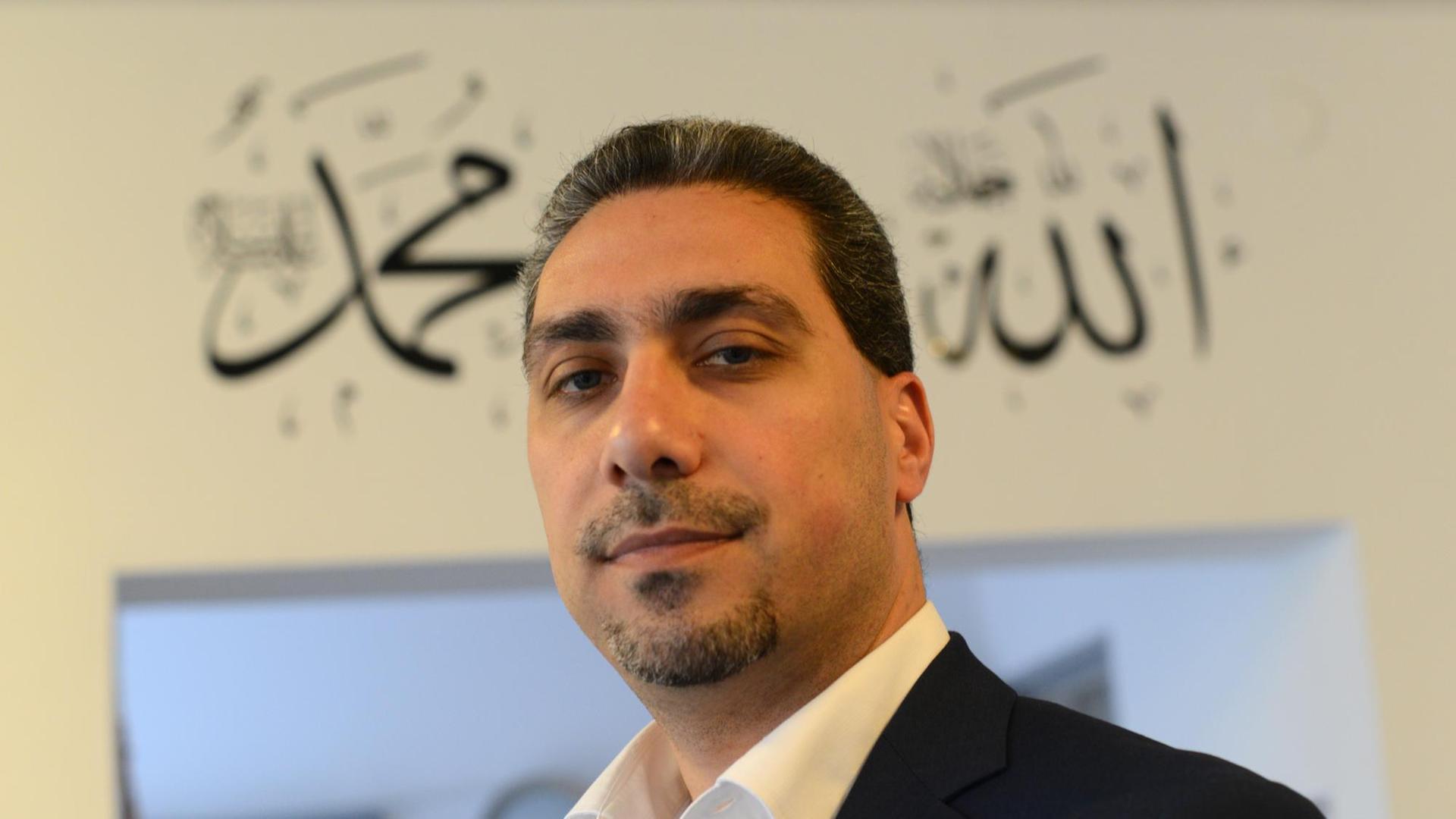 Sadiqu Al-Mousllie im schwarzen Sakko und weißem Hemd vor einem Türeingang mit arabischen Schriftzügen. Er blickt in die Kamera. Sadiqu Al-Mousllie im schwarzen Sakko und weißem Hemd vor einem Türeingang mit arabischen Schriftzügen. Er blickt in die Kamera.