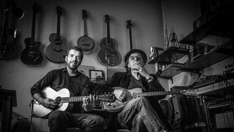Die Gitarristen Danny Dziuk und Antoine Villoutreix sitzen in einem Raum, an dessen Wand viele Gitarren hängen.
