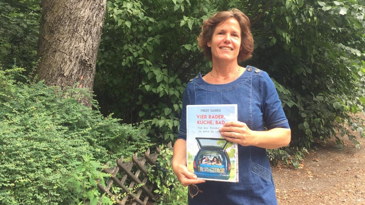 DLF Kultur-Literaturredakteurin Elke Schlinsog. Sie steht in einem Park und hält ein Buch in der Hand: "Vier Räder, Küche, Bad" von Fredy Gareis.