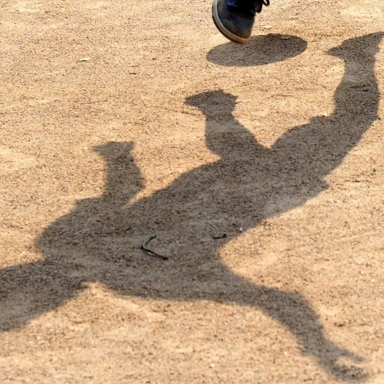 Der Schatten eines Jugendlichen mit einem Fußball zeichnet sich auf einem Bolzplatz ab