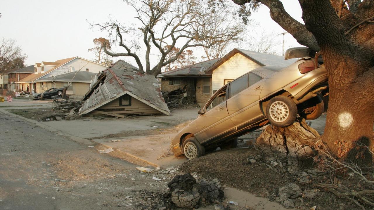 Zu sehen sind ein Auto, das durch Hurrikan Katrina schräg an einen Baum geworfen wurde, sowie abgedeckte Hausdächer auf der Straße.
