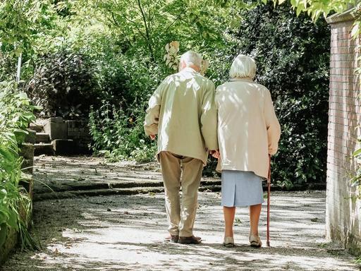 Ein alter Mann und eine alte Frau machen gemeinsam einen Spaziergang. Der Weg ist Bäumen und Büschen flankiert, auf der rechten Seite befindet sich eine Mauer.