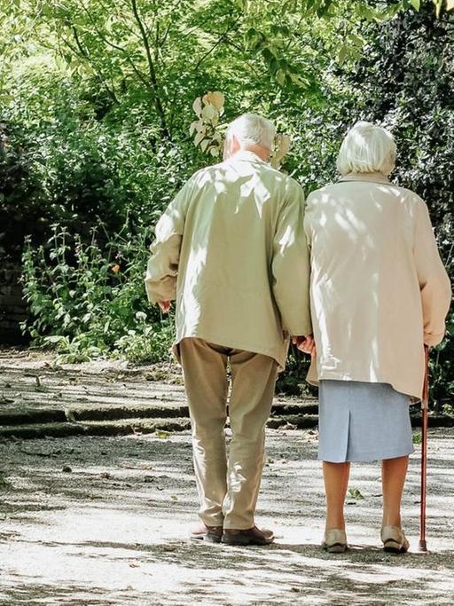 Ein alter Mann und eine alte Frau machen gemeinsam einen Spaziergang. Der Weg ist Bäumen und Büschen flankiert, auf der rechten Seite befindet sich eine Mauer.