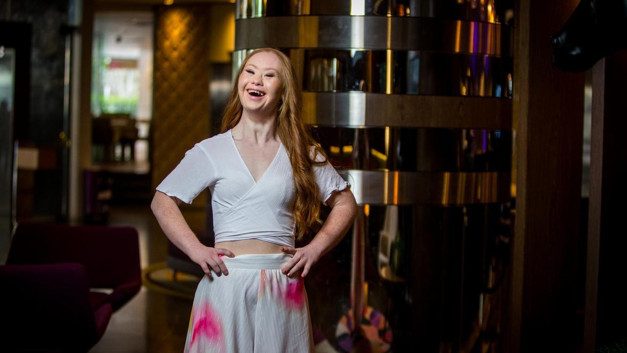 Madeline Stewart trägt ein weißes Oberteil und weißen Rock mit pinken Flecken, hat die Hände in die Seiten gestützt und lacht in die Kamera. Madeline Stewart trägt ein weißes Oberteil und weißen Rock mit pinken Flecken, hat die Hände in die Seiten gestützt und lacht in die Kamera.