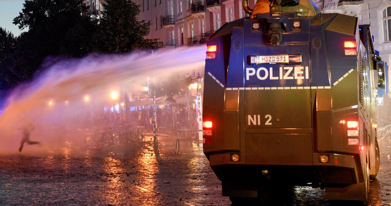 Das Bild zeigt einen Wasserwerfer in der Nacht von hinten, der gerade seitlich Wasser gegen einen Demonstranten verspritzt.