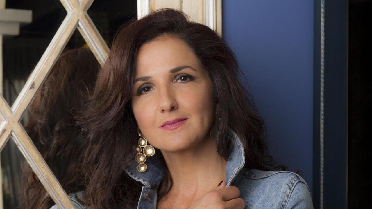 Eine Frau mit langen schwarzen Haaren in Jeans-Jacke steht vor einem Fenster und blickt in die Kamera. Eine Frau mit langen schwarzen Haaren in Jeans-Jacke steht vor einem Fenster und blickt in die Kamera.