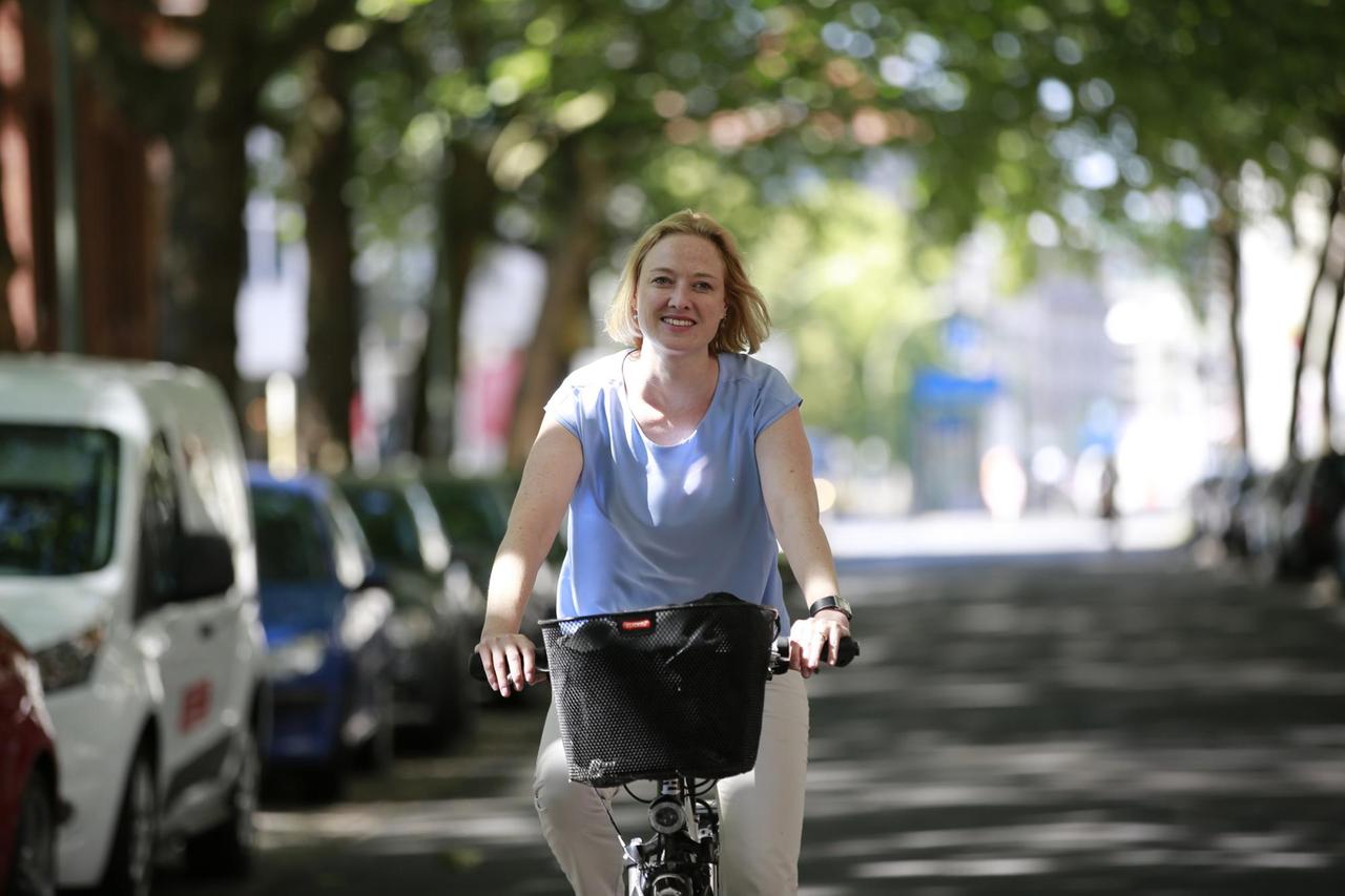 Anika Meenken, Sprecherin des Verkehrsclubs Deutschlands, fährt mit dem Fahrrad durch eine von Bäumen gesäumte Straße und lächelt in die Kamera.