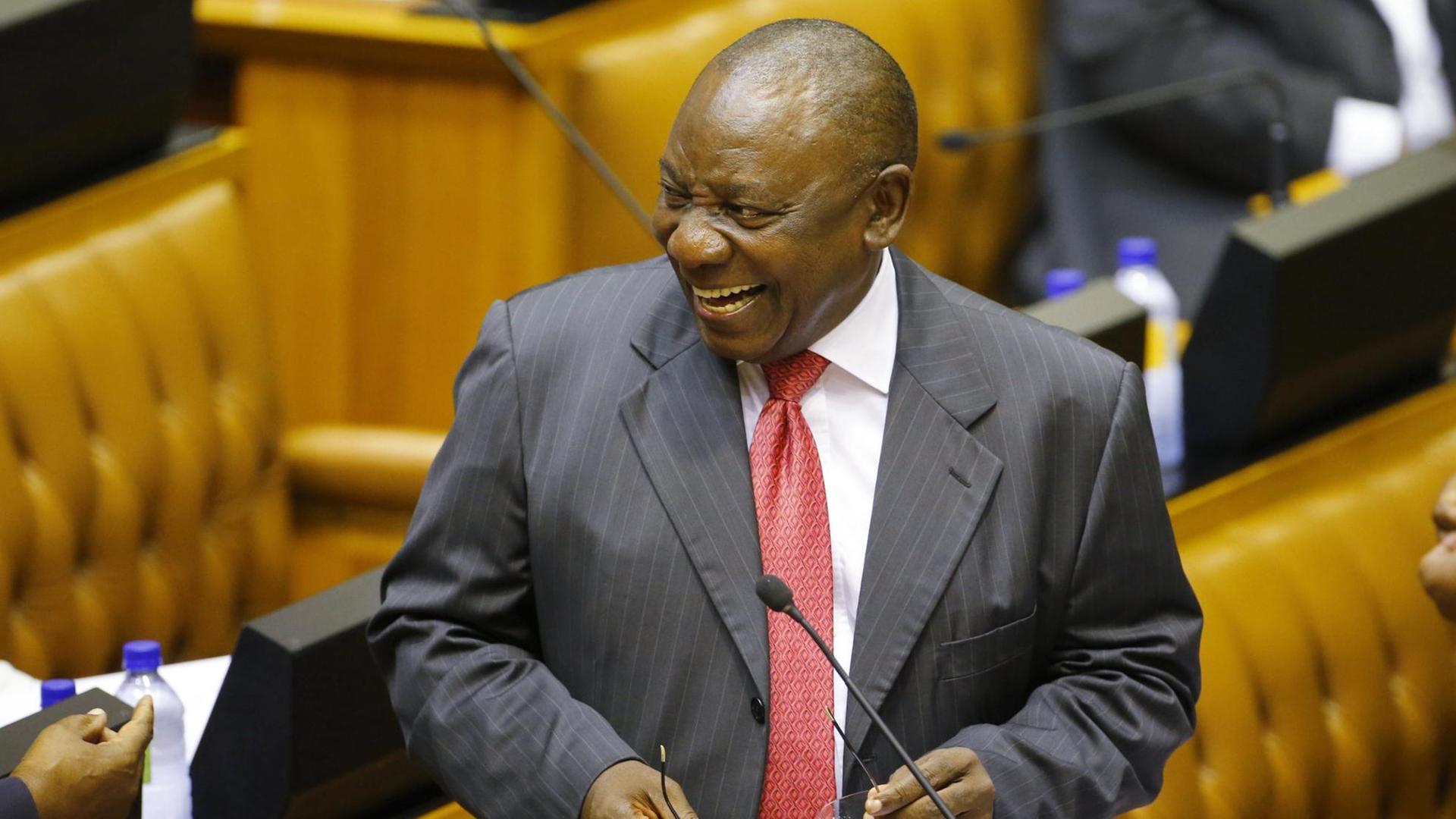 Ramaphosa steht lachend vor einem Mikrofon im Parlament und hält seine Brille in den Händen. Ramaphosa steht lachend vor einem Mikrofon im Parlament und hält seine Brille in den Händen.