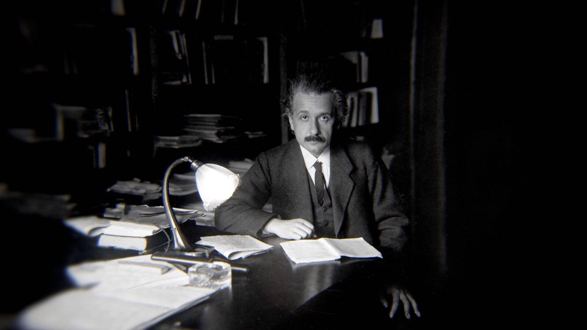 Albert Einstein sitzt an einem Schreibtisch, auf dem eine Lampe brennt, im Hintergrund Bücherregale Albert Einstein sitzt an einem Schreibtisch, auf dem eine Lampe brennt, im Hintergrund Bücherregale