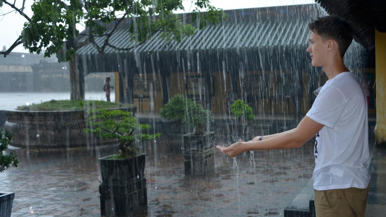 Ein junger Mann steht unter einem Vordach und hält die Hand unter den Regen.