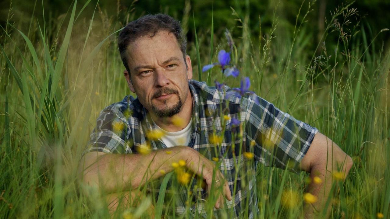 Der Autor des Buchs Wiese sitzt im Gras und blickt auf gelbe und blaue Blüten.
