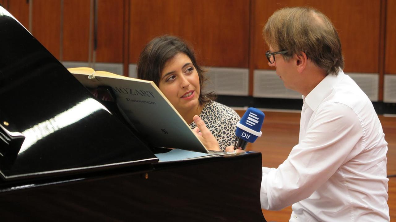 Eine Pianistin und ein Moderator sitzen an einem Flügel in einem Konzertsaal und sprechen miteinander. Eine Pianistin und ein Moderator sitzen an einem Flügel in einem Konzertsaal und sprechen miteinander.