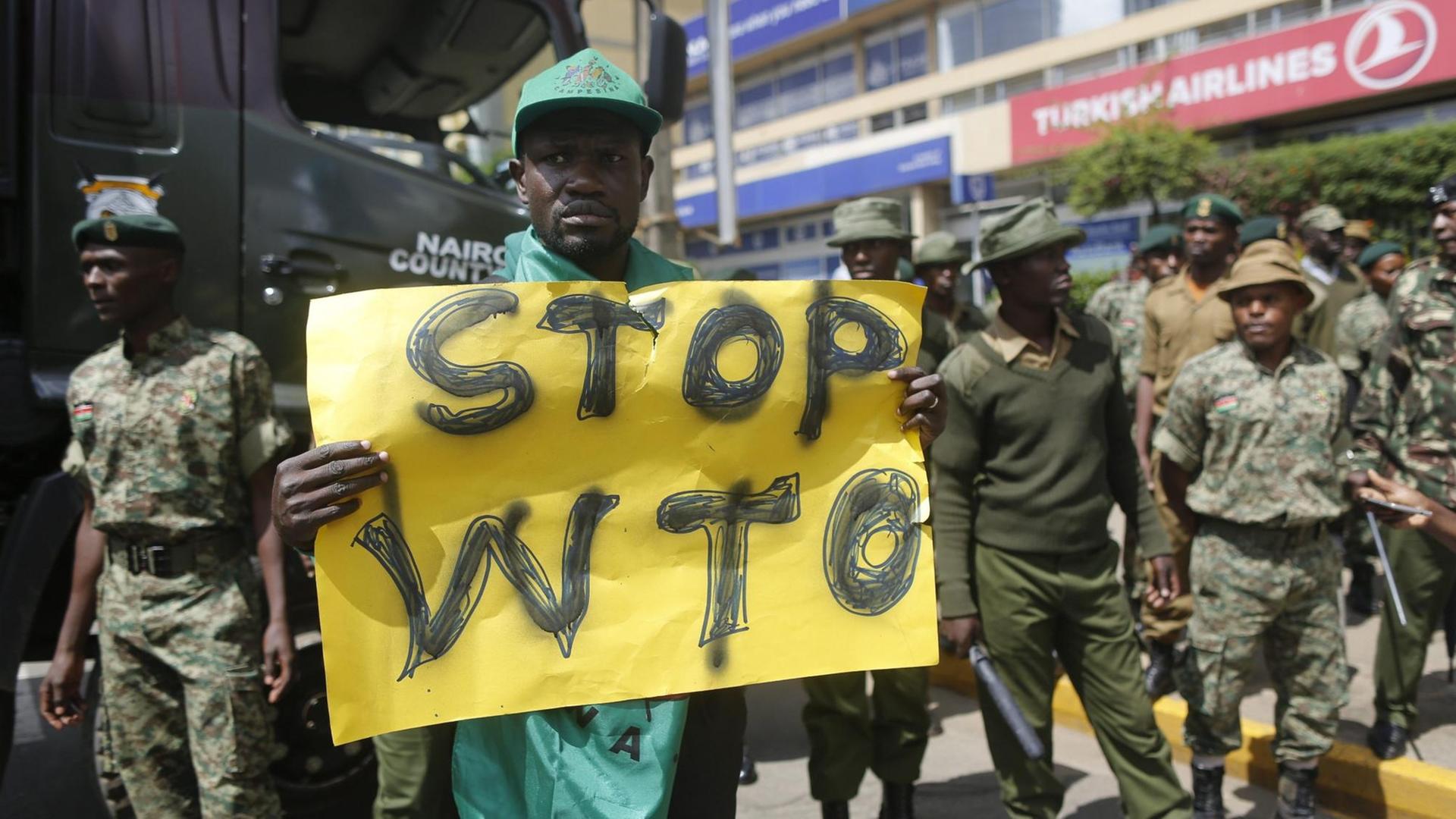 Ein kenianischer Aktivist hält ein Transparent hoch, auf dem "Stop WTO" zu lesen ist. Um ihn herum stehen Sicherheitsleute. Ein kenianischer Aktivist hält ein Transparent hoch, auf dem "Stop WTO" zu lesen ist. Um ihn herum stehen Sicherheitsleute.