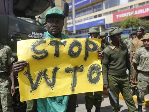 Ein kenianischer Aktivist hält ein Transparent hoch, auf dem "Stop WTO" zu lesen ist. Um ihn herum stehen Sicherheitsleute.