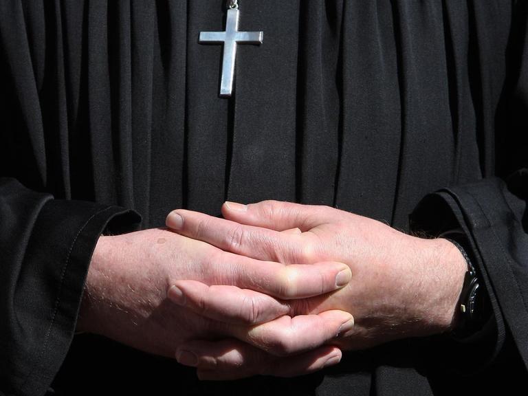 Ein Pfarrer im schwarzen Talar mit gefalteten Händen, auf der Brust ein silbernes Kreuz.