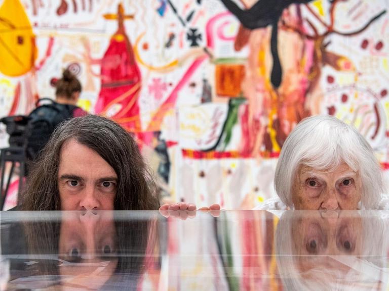 Brigitte und Jonathan Meese (Maler und Aktionskünstler) spiegeln sich im Ausstellungsraum in einem Schaukasten. Im Hintergrund sind Gemälde von Jonathan Meese zu sehen.