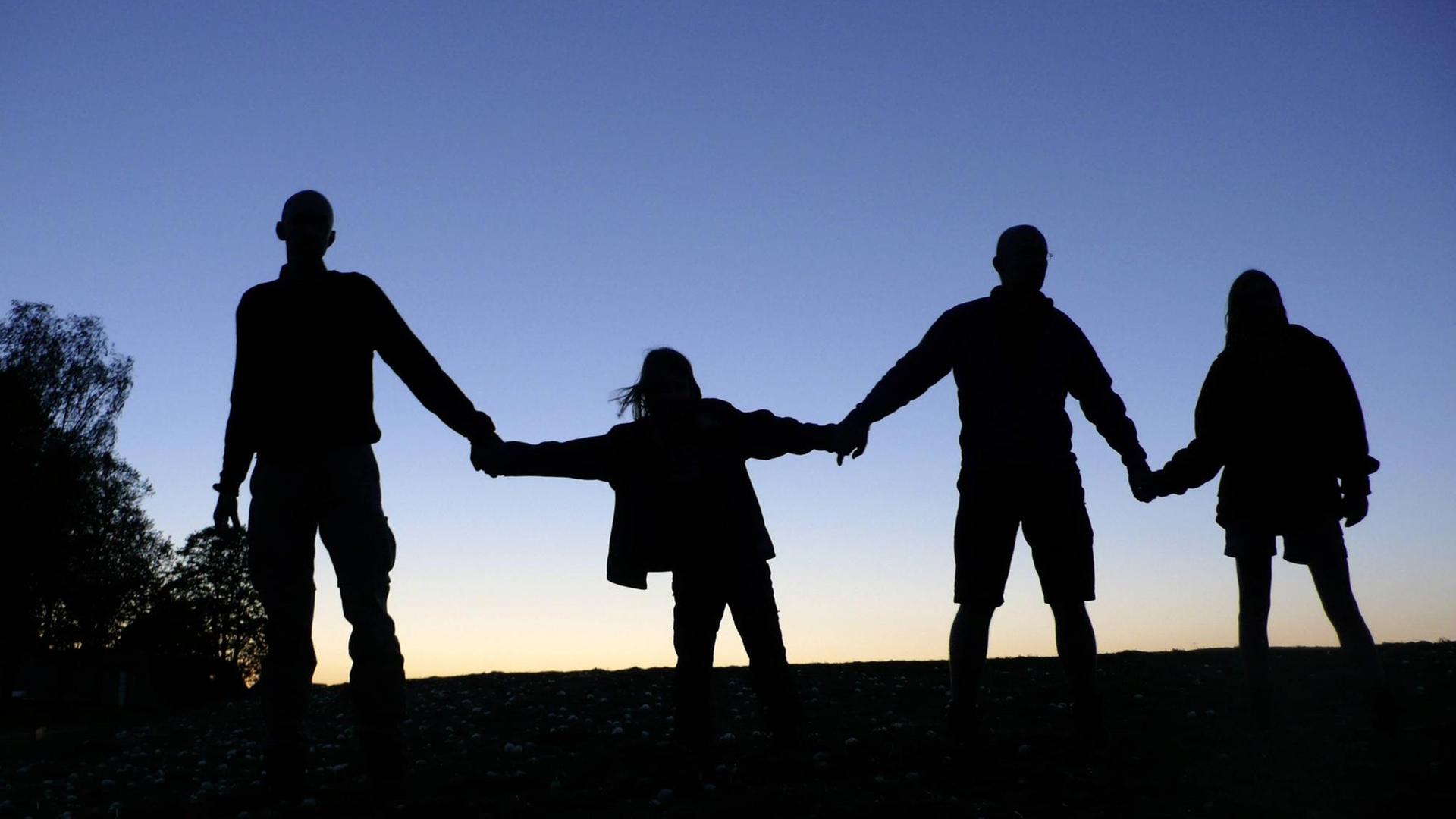 Drei Erwachsene und ein Kind halten sich die Hände, sie sind als Schattenriss vor dem dämmernden Himmel zu sehen. (Symbolbild gesellschaftlicher Zusammenhalt)
