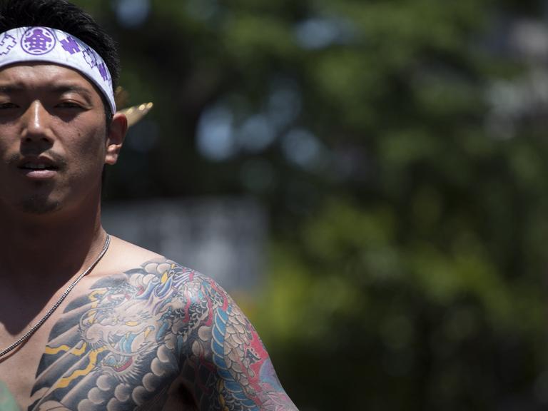 Ein junger Japaner zeigt seinen großflächig tätowierten Oberkörper. Er hat sich ein weißes Band mit lila Zeichen um den Kopf gebunden.