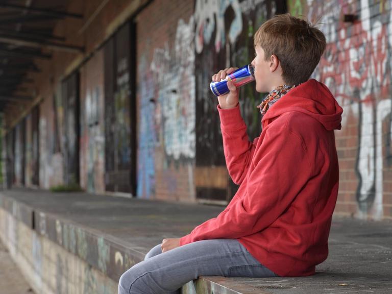 Ein Jugendlicher sitzt auf einer Steinmauer und trinkt einen Energy Drink.