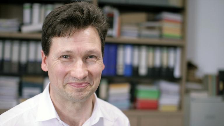 Dominik Burkard, Professor für Kirchengeschichte an der Universität Würzburg. Ein Mann mit kurzen dunklen Haaren steht in einem Büro und lächelt in die Kamera.