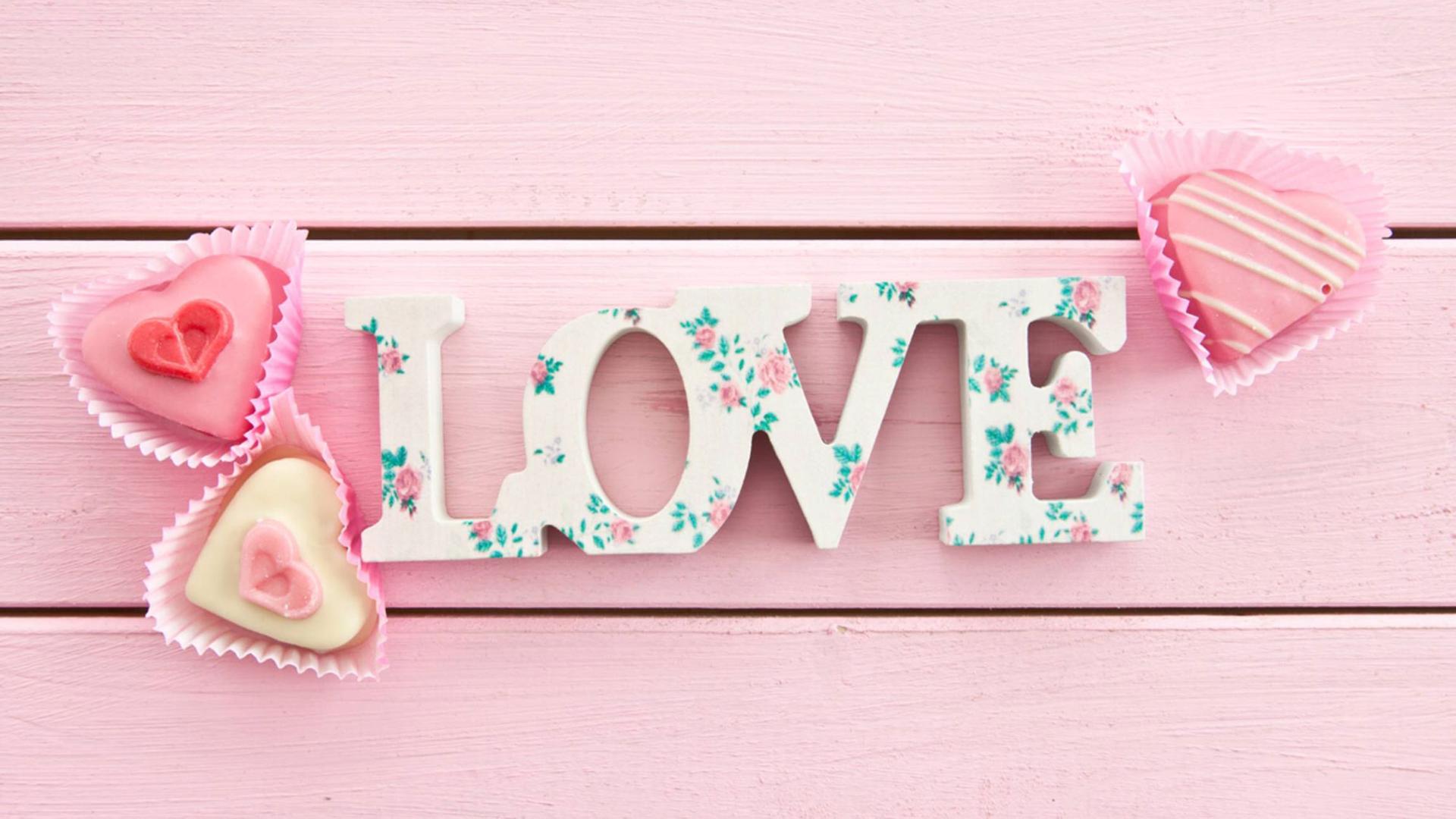 Der Schriftzug "Liebe" liegt auf rosa gestrichenem Holz, daneben drei rosafarbene Törtchen in Herzform.