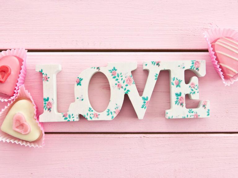 Der Schriftzug "Liebe" liegt auf rosa gestrichenem Holz, daneben drei rosafarbene Törtchen in Herzform.