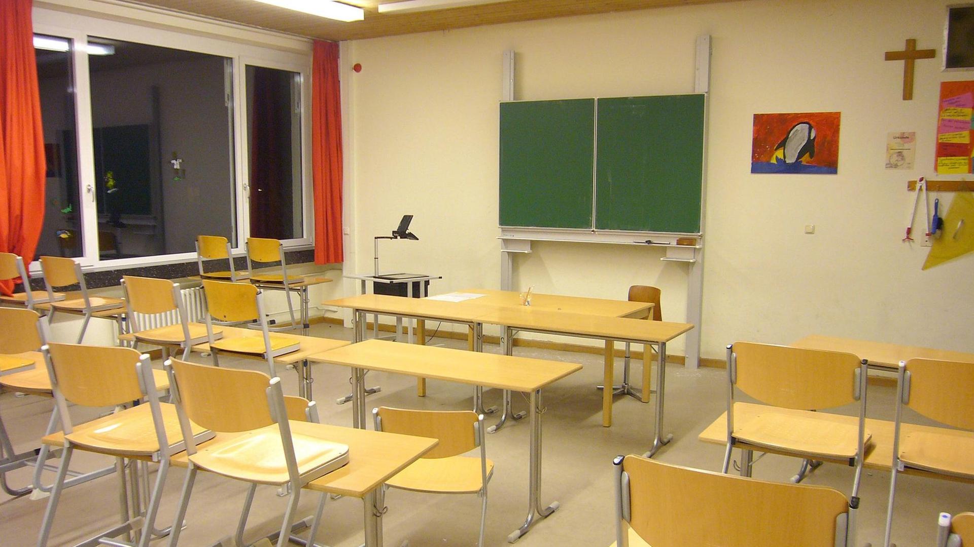 Ein leeres Klassenzimmer. Die Tafel ist abgewischt und zugeklappt. Die Stühle aus hellbraunem Holzfurnier stehen auf den Tischen.