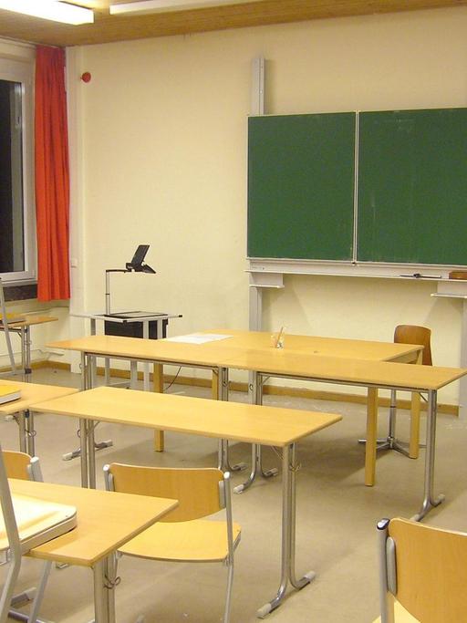 Ein leeres Klassenzimmer. Die Tafel ist abgewischt und zugeklappt. Die Stühle aus hellbraunem Holzfurnier stehen auf den Tischen.
