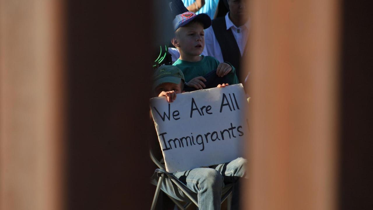 Blick durch die Stäbe der Grenzmauer zu Mexico. Ein Junge hält ein Transparent mit der Aufschrift "Wir sind alle Migranten" in den Händen.