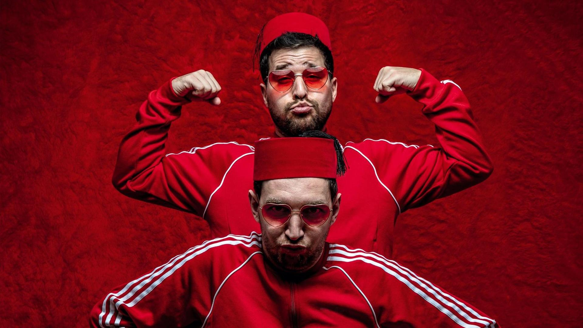Zu sehen sind zwei Männer, die hintereinanderstehen. Sie tragen einen Fes auf dem Kopf und rote Adidas-Trainingsanzüge. Zu sehen sind zwei Männer, die hintereinanderstehen. Sie tragen einen Fes auf dem Kopf und rote Adidas-Trainingsanzüge.