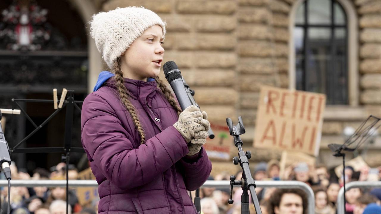 Die schwedische Klimaaktivistin Greta Thunberg spricht auf einer Bühne in ein Mikrofon. Im Hintergrund stehen viele demonstrierende Menschen und halten Banner in die Luft. 