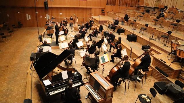 Mitglieder eines Orchesters sitzen breit verteilt in einem Konzertsaal und musizieren vor leeren Rängen.
