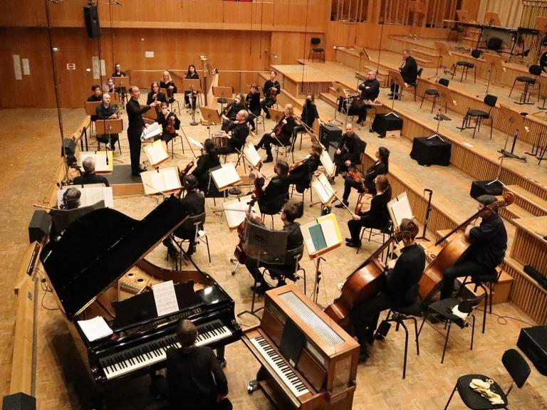 Mitglieder eines Orchesters sitzen breit verteilt in einem Konzertsaal und musizieren vor leeren Rängen.