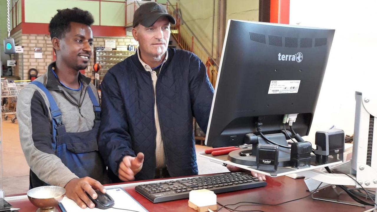 Ein Mann mittleren Alters mit Ziegenbärtchen erklärt einem eritreischen jungen Mann ein technisches Detail am Computer Ein Mann mittleren Alters mit Ziegenbärtchen erklärt einem eritreischen jungen Mann ein technisches Detail am Computer