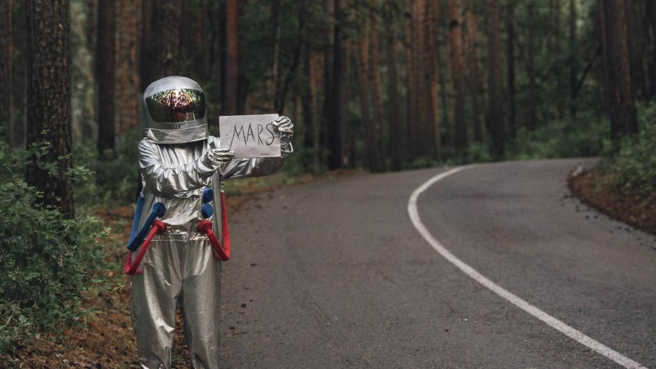 Ein als Astronaut verkleideter Tramper steht am Strassenrand und hält ein Pappschild hoch auf dem "Mars" steht. Ein als Astronaut verkleideter Tramper steht am Strassenrand und hält ein Pappschild hoch auf dem "Mars" steht.