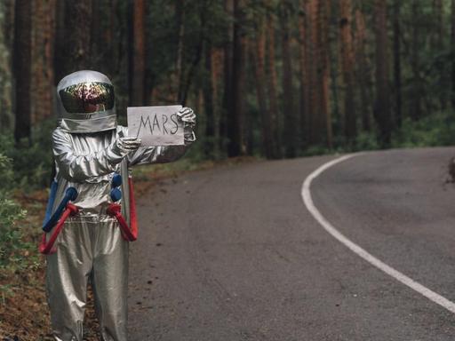 Ein als Astronaut verkleideter Tramper steht am Strassenrand und hält ein Pappschild hoch auf dem "Mars" steht.