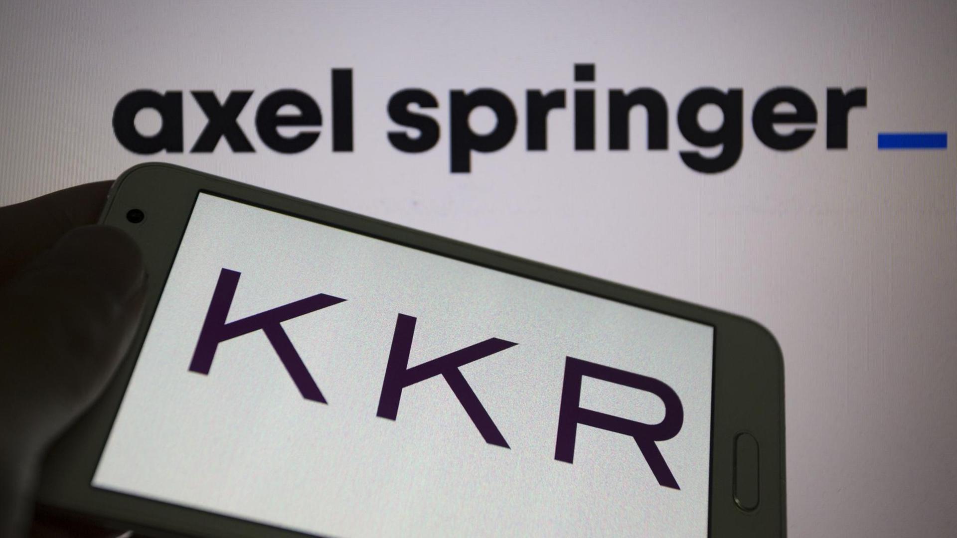 Das Logo von KKR (Kohlberg Kravis Roberts & Co.) ist auf einem Smartphone zu sehen. Im Hintergrund ist auf einer Wand das Logo von Axel Springer zu sehen.