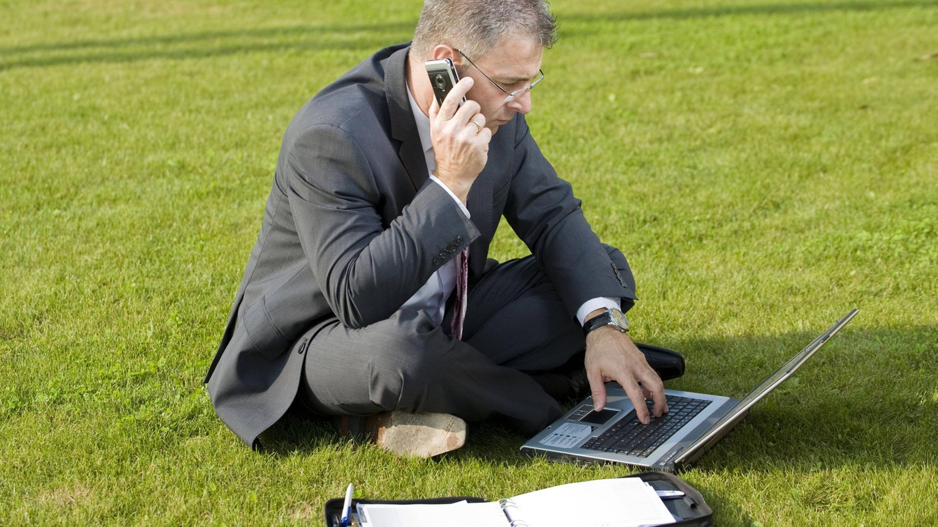 Ein Geschäftsmann sitzt im Anzug auf einer Wiese und telefoniert mit dem Handy, vor ihm Laptop und Ringbuch. Ein Geschäftsmann sitzt im Anzug auf einer Wiese und telefoniert mit dem Handy, vor ihm Laptop und Ringbuch.