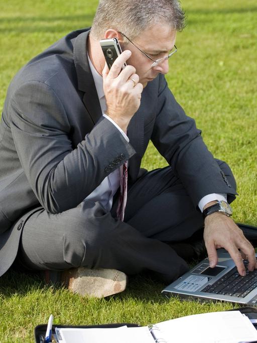 Ein Geschäftsmann sitzt im Anzug auf einer Wiese und telefoniert mit dem Handy, vor ihm Laptop und Ringbuch.