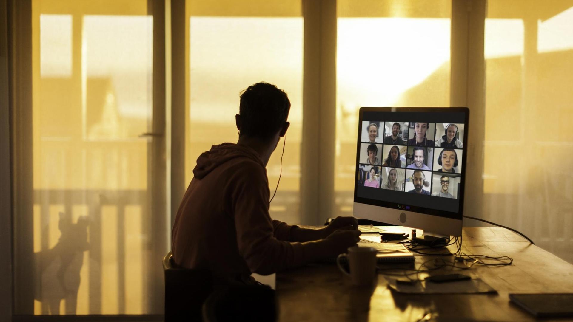 Ein junger Mann sitzt zu Hause vor einem Bildschirm mit Teilnehmern eines digitalen Treffens.